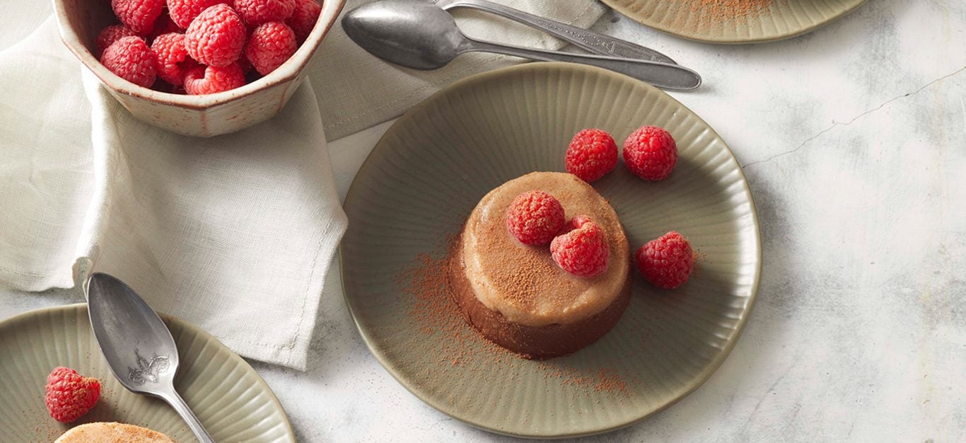 Date-Sweetened Chocolate Vegan panna Cotta - Light caramel colored panna cotta drizzled with fresh raspberries, sitting on a gray plate, with a bowl of raspberries on the side