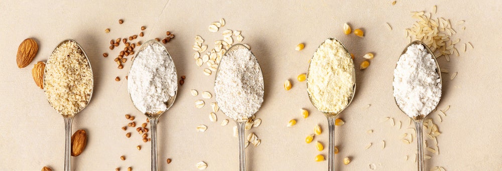 five spoons lying side by side, each with a different type of gluten-free flour in it - almond flour, buckwheat flour, oat flour, corn flour, brown rice flour