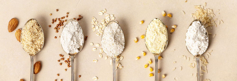 five spoons lying side by side, each with a different type of gluten-free flour in it - almond flour, buckwheat flour, oat flour, corn flour, brown rice flour