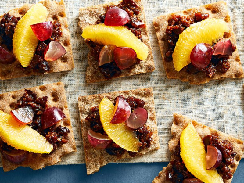 Four Grape and Fig Snacks on whole grain crackers, on a rough linen fabric on a blue table