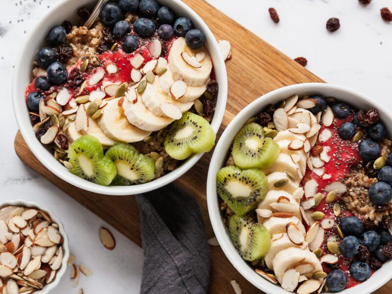 Two bowls of Berry-Licious Overnight Oatmeal next to smaller bowls of raisins, slivered almonds, and chia seeds