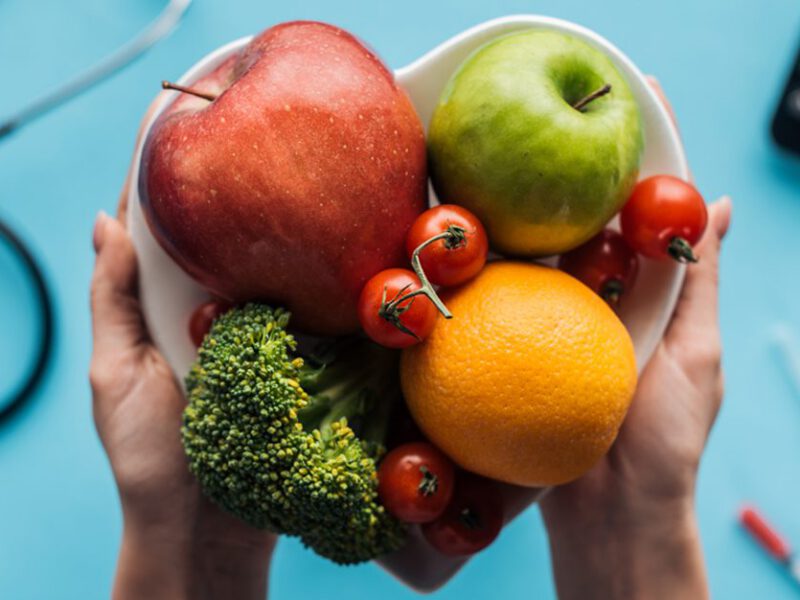 Two hands hold a heart-shaped bowl filled with an orange, two apples, broccoli, and cherry tomatoes. In the background, which is out of focus, are a stethoscope and blood glucose monitor