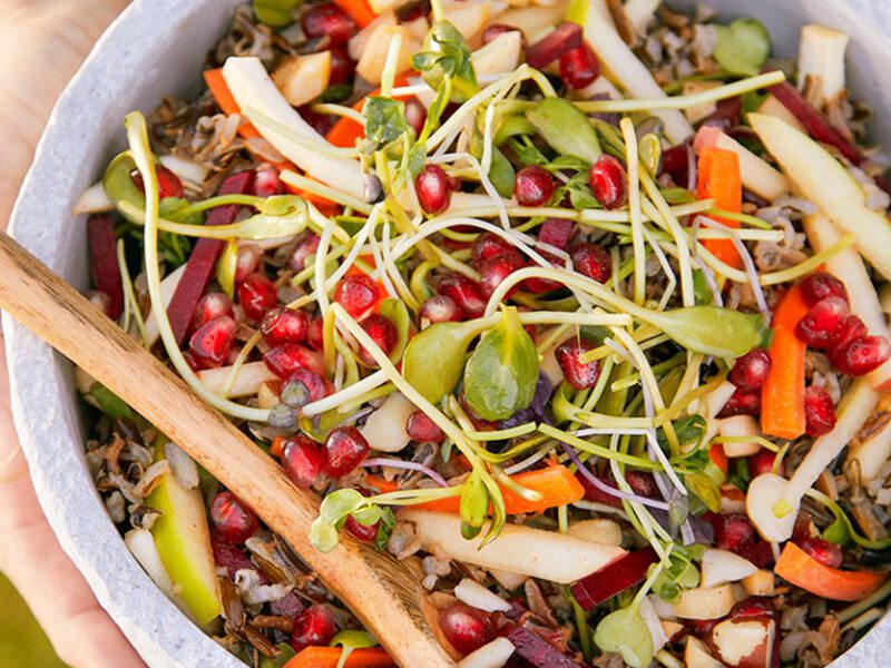 Hands holding a white marble bowl of Pomegranate Wild Rice Salad, with a wooden serving spoon in the bowl