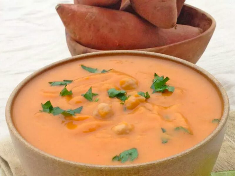 A bowl of Spicy Sweet Potato Bisque with Chickpeas in the foreground, with a bowl of raw sweet potatoes just behind it