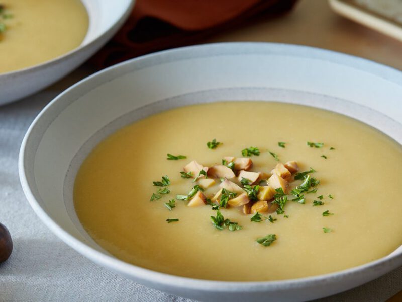 A wide-rimmed pale grey bowl of Winter Potato Leek Soup. Next to the bowl are chestnuts and dried rosemary