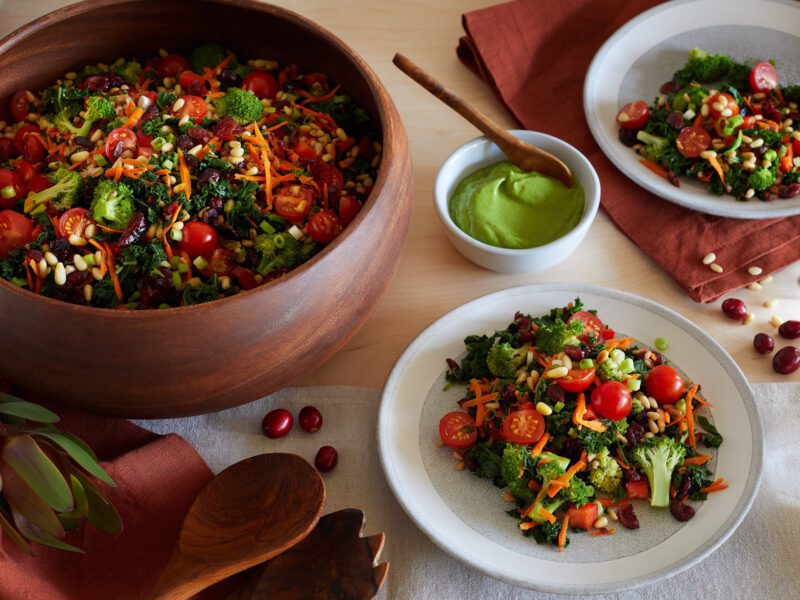 A large bowl of kale salad set alongside a plate with kale salad and a bowl with green avocado dressing
