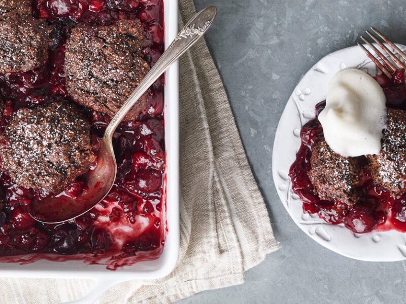 Chocolate Cherry Cobbler shown in a baking dish with serving spoon, with one serving on a plate shown with whipped cream topping