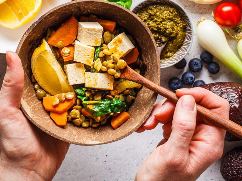 Man holds up bowl with colorful array of vegetables, tofu, grains, and lemon wedge, while more fruits and veggies are on the table below