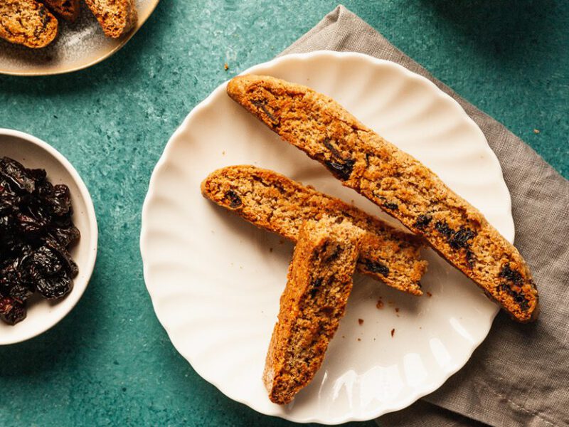 Three slices of Cranberry-Orange Biscotti on a white plate next to a small bowl of dried cranberries