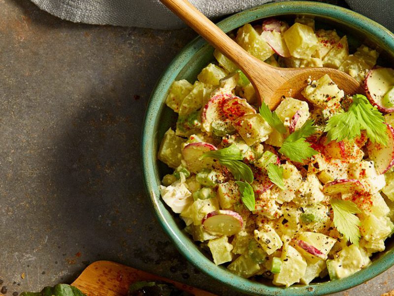 Vegan Egg Salad - Diced potatoes, celery, and other veggies and herbs shown tossed together for a vegan egg salad, in a green bowl on a concrete table
