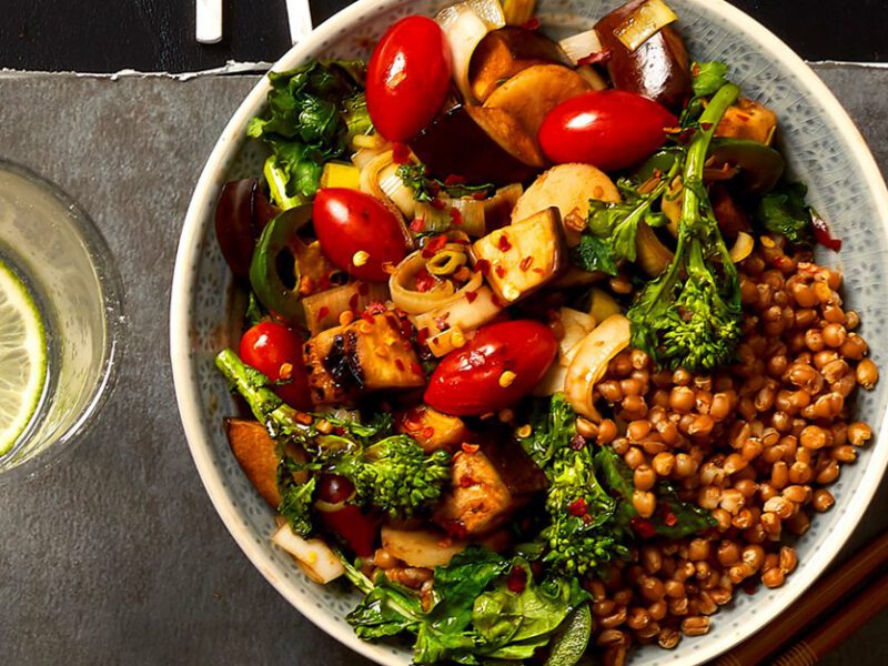 An inviting bowl of Szechwan Stir-Fry featuring wheat berries, cherry tomatoes, broccolini, and water chestnuts on a grey counter with a glass of lime water and chopsticks beside it.