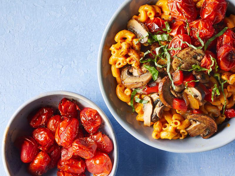 Vegan Mac and Cheese with Queso against a blue background next to a small bowl of roasted cherry tomatoes
