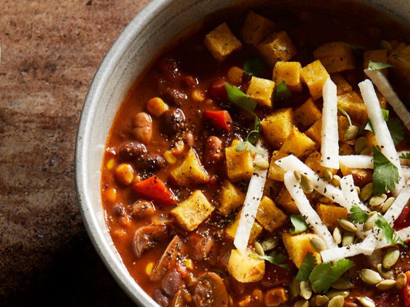 A pot of deep red mole-style vegan chili topped with corn and strips of jicama