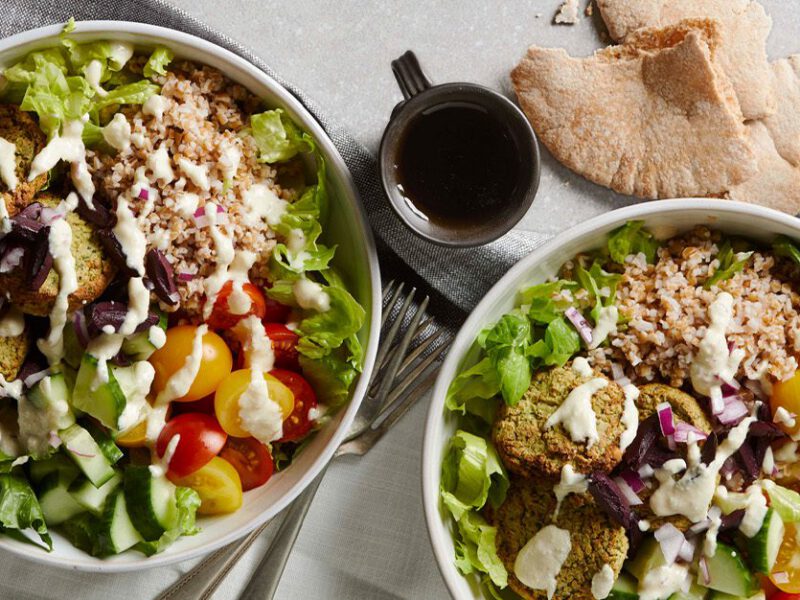 Two bowls loaded with a colorful mix of chopped lettuce, cucumbers, tomatoes over a pile of bulgur grains, topped with air-fryer falafel and drizzled with sauce