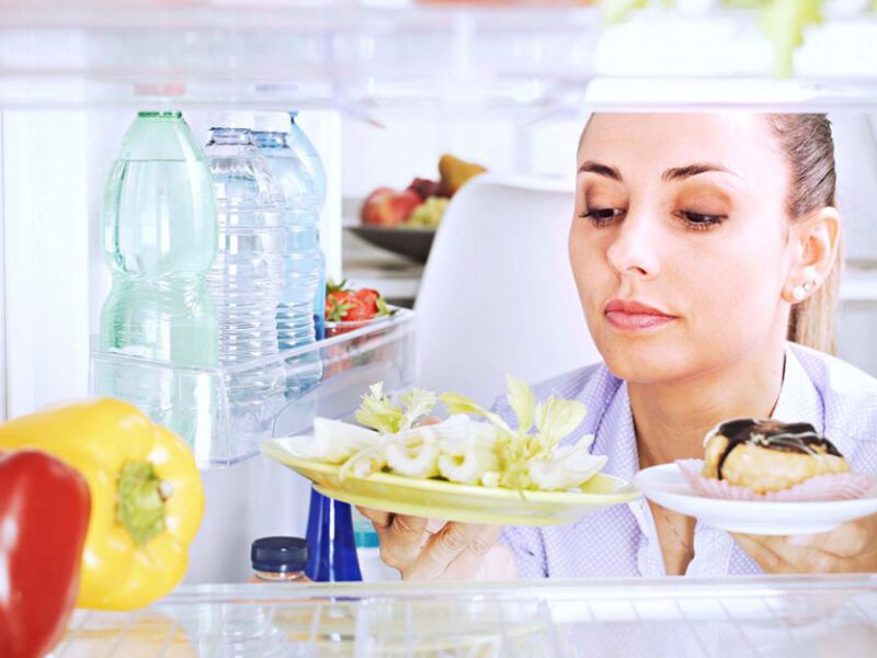 Photo taken from inside a refrigerator, with a woman looking in and examining a donut and vegetables