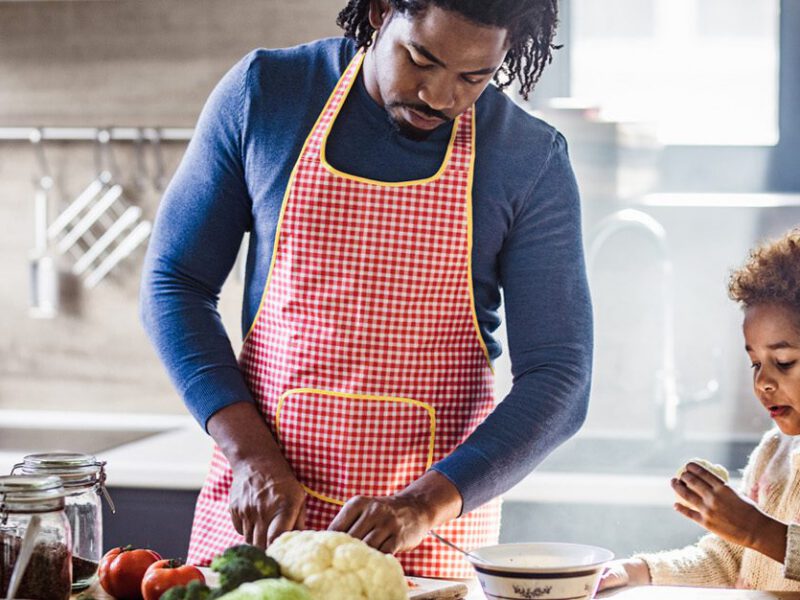 Black father and daughter in kitchen chopping vegetables, with daughter examining a piece of vegetable - teaching children about cultures through food