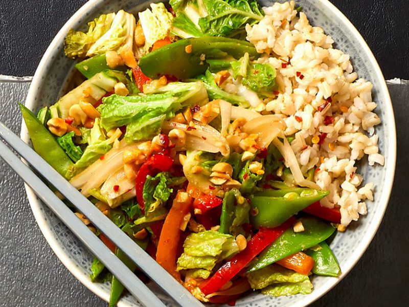 peanut sauce stir-fry - array of shredded cabbage, snap peas, and bell peppers tossed together and served over a bed of brown rice, shown on a gray table next to a glass of sparkling water, with chopsticks on the side