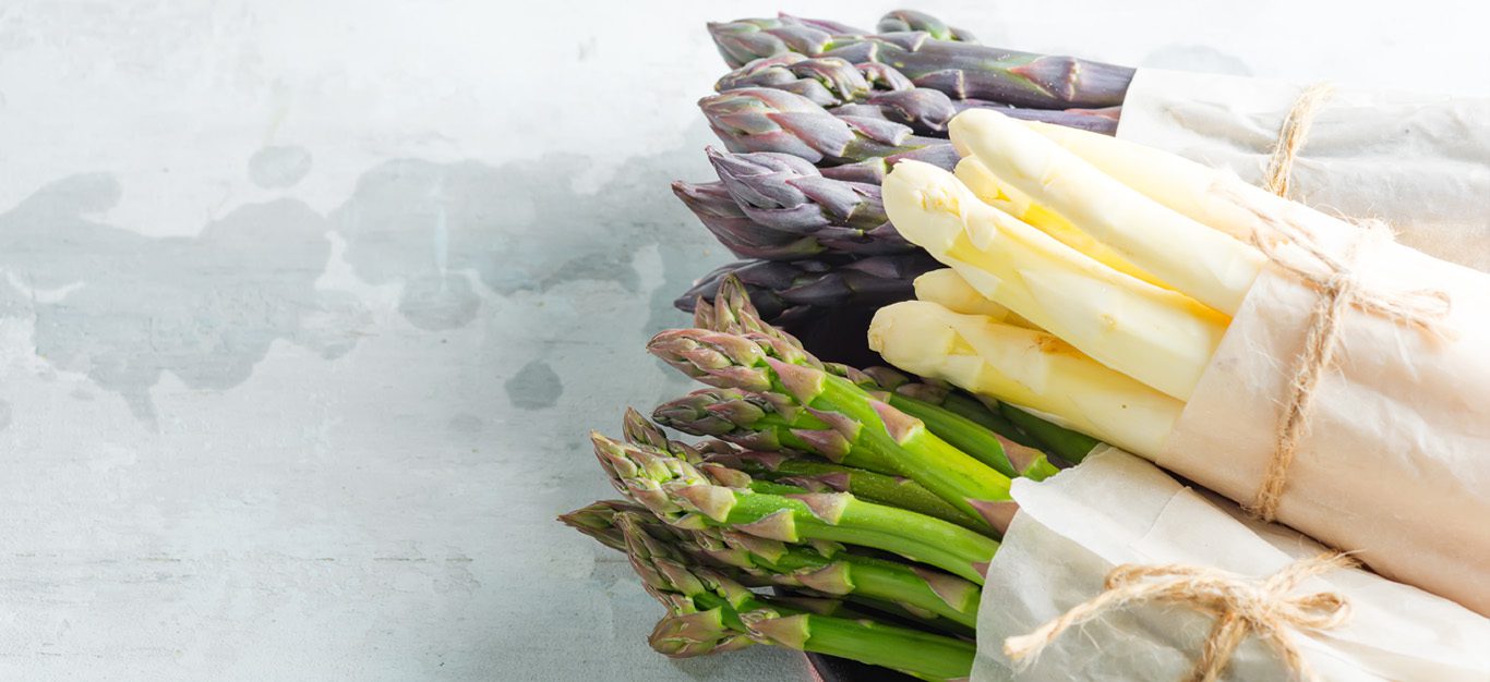 Three bundles of asparagus, green, white, and purple, on a white table