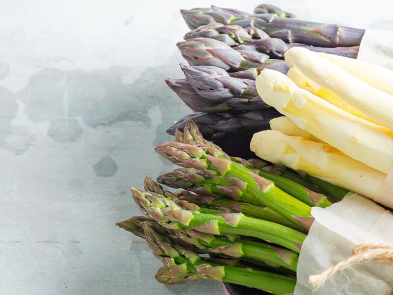 Three bundles of asparagus, green, white, and purple, on a white table