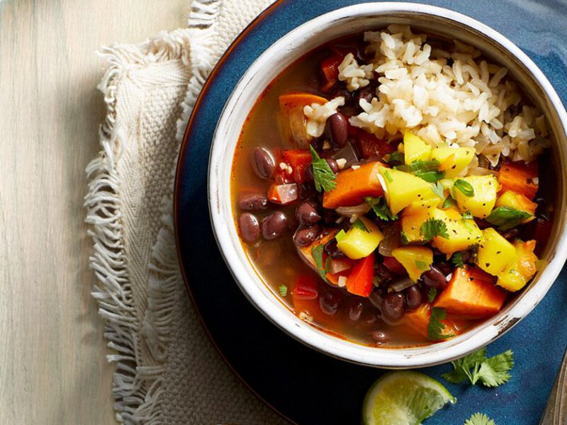 A Caribbean-inspired black bean soup, served over brown rice, topped with diced mango relish, in a white bowl atop a blue plate next to a spoon