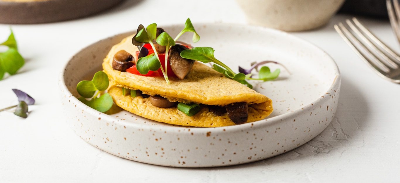 photo of a folded vegan chickpea omelette with mushrooms visible inside, topped with diced tomatoes and garnished with microgreens, on a white plate