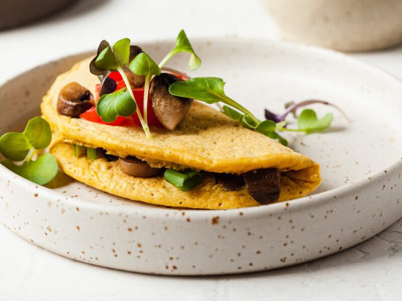 photo of a folded vegan chickpea omelette with mushrooms visible inside, topped with diced tomatoes and garnished with microgreens, on a white plate