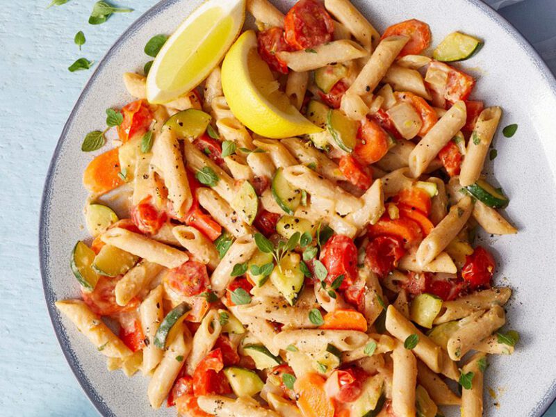 Lemon Hummus Pasta: Penne pasta tossed with a creamy hummus-based dressing, tossed with chopped red bell pepper. Served on a white plate, with lemon wedges on the side.