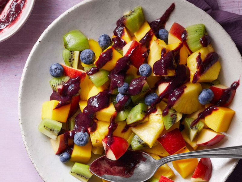 blueberry balsamic glaze sauce shown drizzled over a colorful array of fruit including kiwis and mango, on a purple table