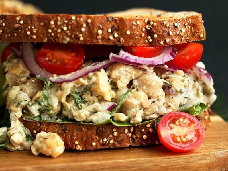 A close-up shot of a vegan tuna salad sandwich with two slices of whole grain bread, with the filling featuring chickpeas, onions, and cherry tomatoes and a vegan dressing
