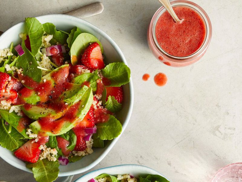 A colorful salad with sliced strawberries, avocado, and greens mixed together and drizzled with a bright red strawberry dressing, with a jar of strawberry dressing on the side