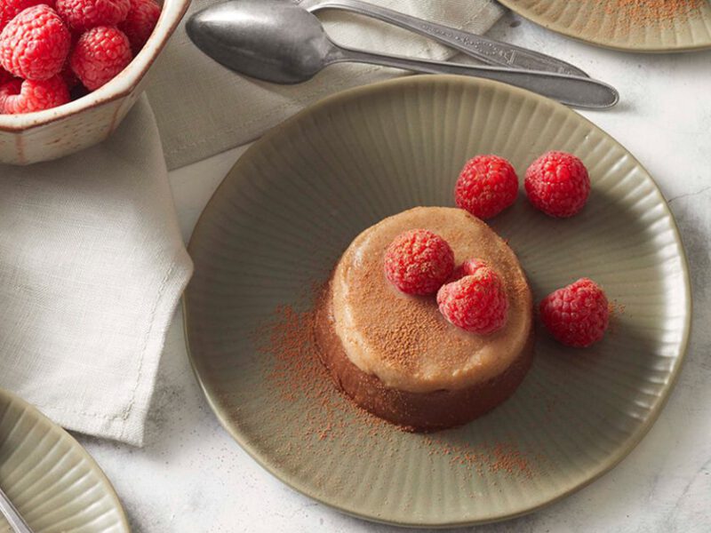 Date-Sweetened Chocolate Vegan panna Cotta - Light caramel colored panna cotta drizzled with fresh raspberries, sitting on a gray plate, with a bowl of raspberries on the side