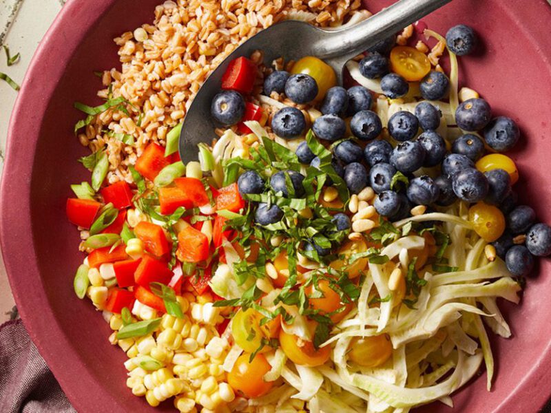 Blueberry Farro Grain Bowl - Colorful array of diced bell pepper tomatoes, fresh herbs, and corn, with farro and blueberries, in a reddish pink bowl with a glass of water beside