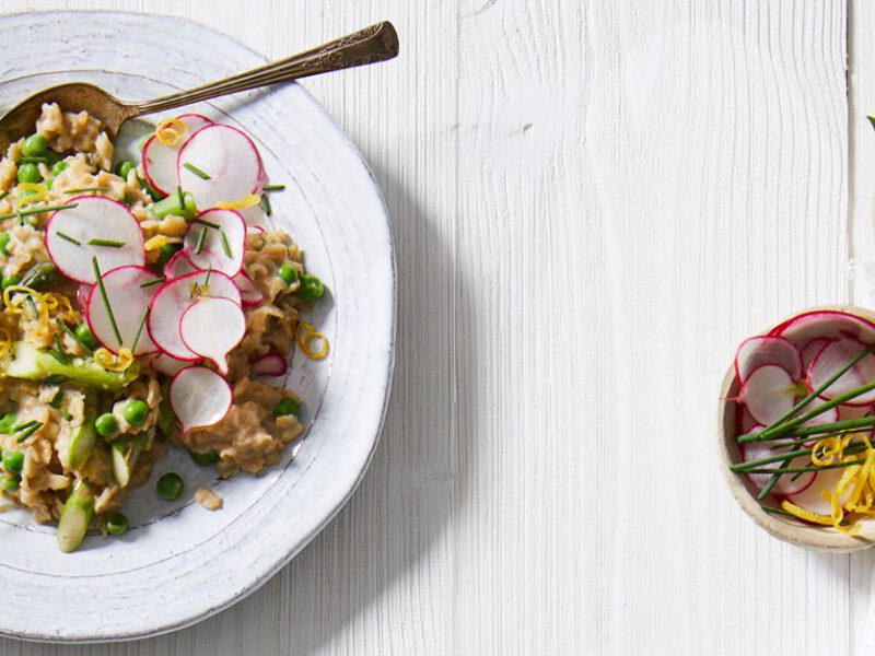 A bowl of savory homemade vegan oatmeal studded with asparagus and peas, and topped with lemon wedges and radish slices, on a white wooden table with a bowl of additional garnishes on the side