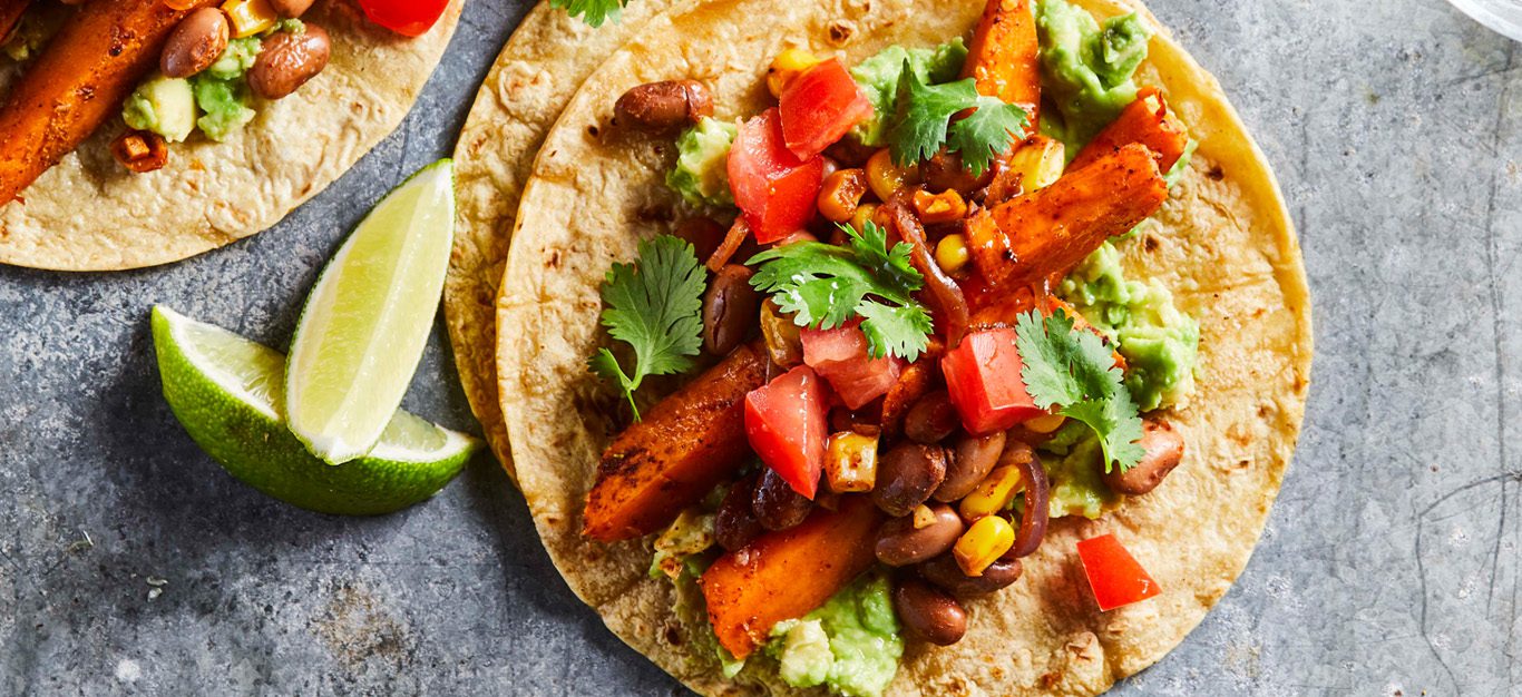 Spiced Sweet Potato Tacos: Vegan tacos shown from above on a gray table, open tortillas topped with sweet potato, peppers, and avocado