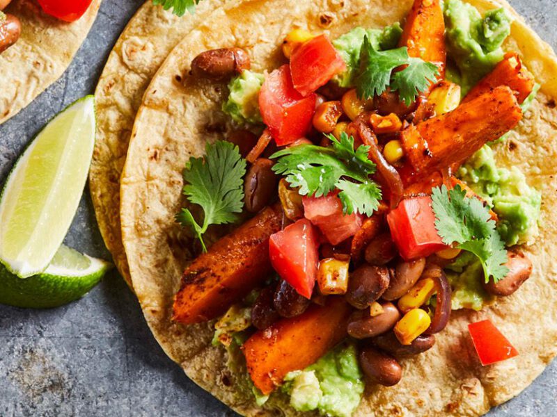 Spiced Sweet Potato Tacos: Vegan tacos shown from above on a gray table, open tortillas topped with sweet potato, peppers, and avocado