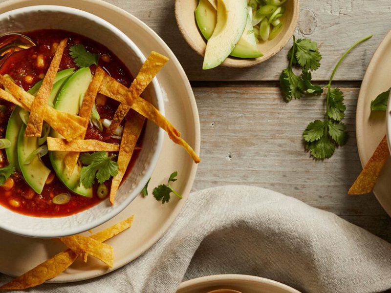 Two bowls of vegan tortilla soup topped with avocado and crunchy tortilla strips, shown on a red table with garnishes between them