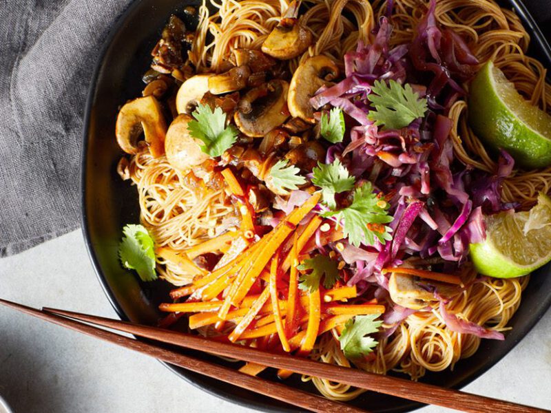 Pancit with Vegetables - Plant-based take on a Filipino dish. Thin rice noodles in a bowl with mushrooms and shredded carrots, with lime wedges and chopsticks on the side