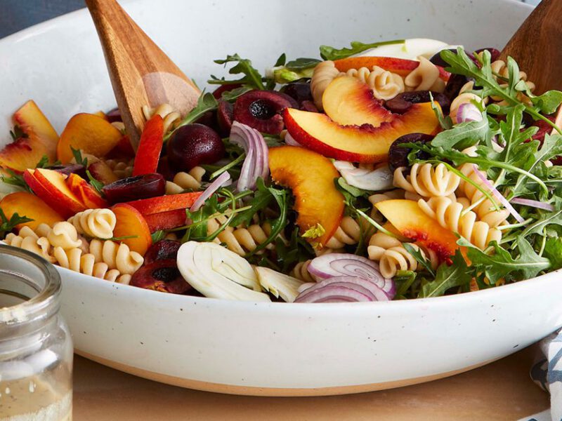 Stone Fruit Pasta Salad in white bowl with wooden salad tossers on a light wooden table