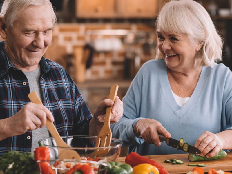 Elderly couple smiles at each other as they chop vegetables to make a salad