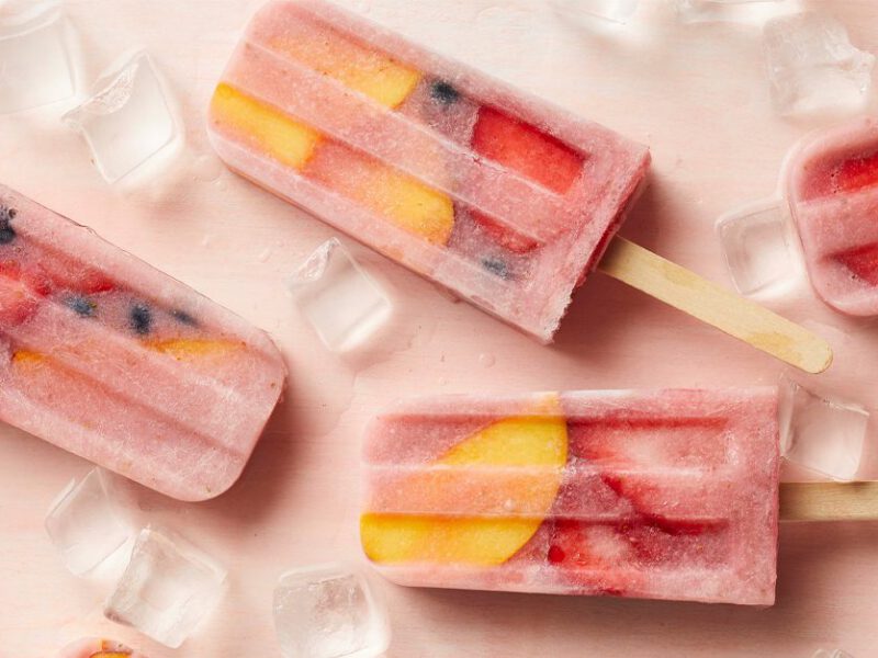 Strawberry, blueberry, and nectarine popsicles on a pink background surrounded by ice cubes