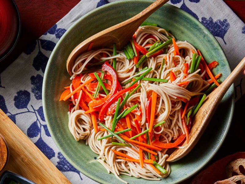 Soba Noodle Salad in a light blue bowl being tossed by a wooden fork and spoon