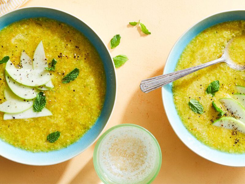 Sunshine Gazpacho topepd with apple slices and mint in blue bowls on a peach colored tabletop