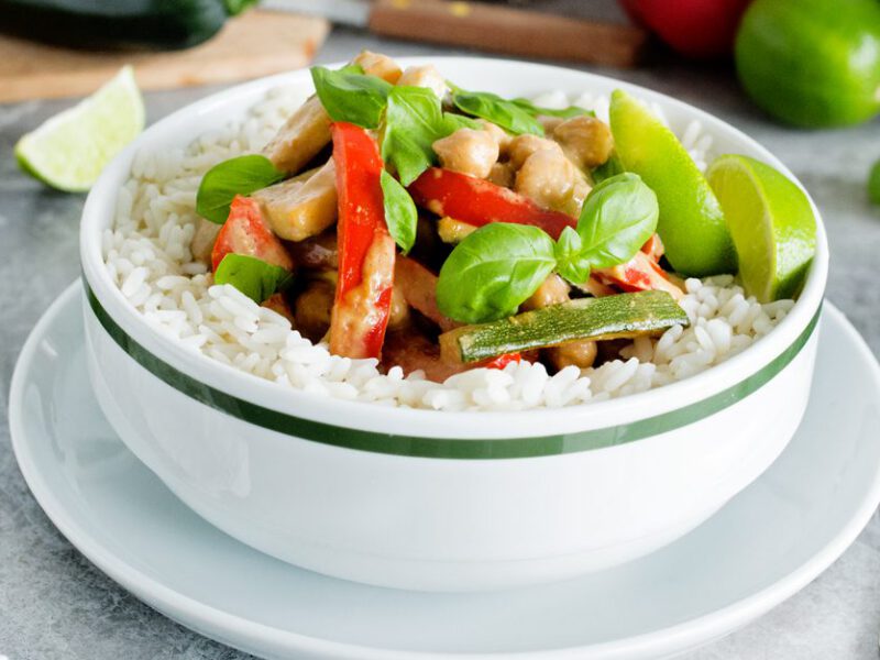 Thai Chickpea-Almond Curry in a white bowl surrounded by fresh limes wedges, basil, and ginger root