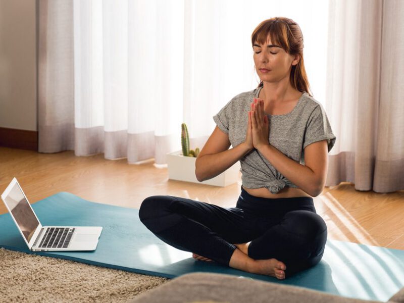 Woman sitting on yoga mat in her home meditating with an open laptop on the floor