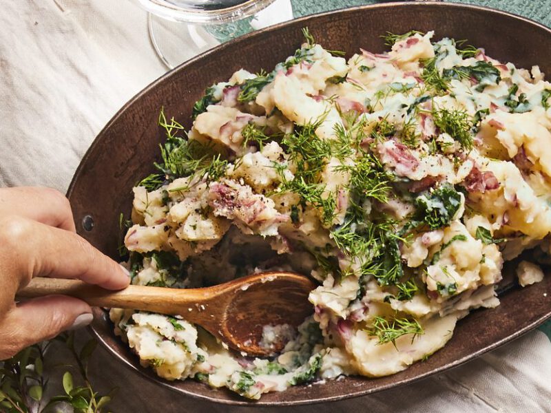 Mashed Potatoes with Kale and fresh dill in a metal serving dish being stirred by a woman's hand