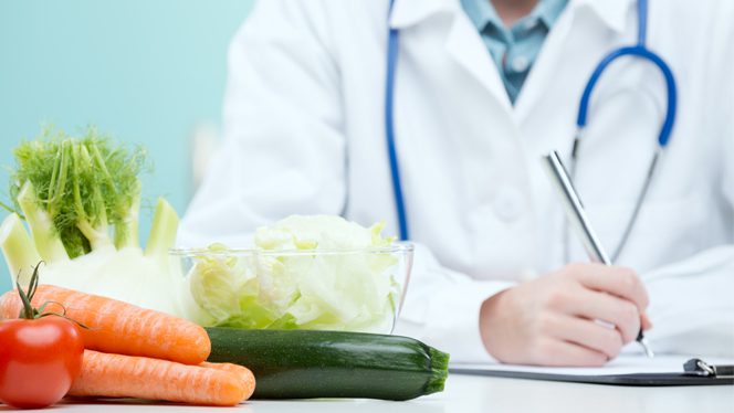 A doctor with a white coat and stethoscope writes notes with vegetables on the table in the foreground