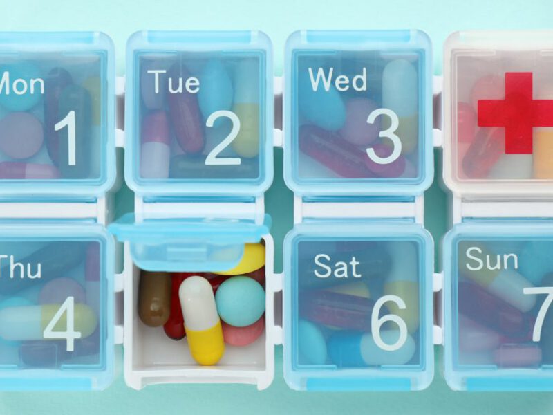 A weekly medication holder is stuffed full of pills against a blue background