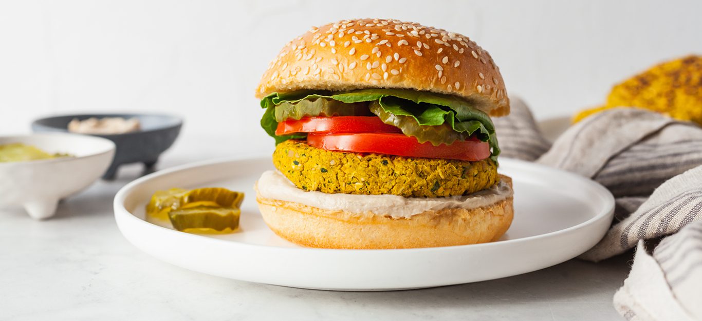 Falafel burger shown in profile, with a bun, falafel patty, and lettuce and tomato toppings, with pickles on the side