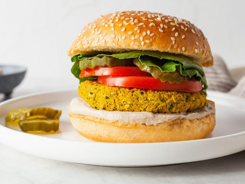 Falafel burger shown in profile, with a bun, falafel patty, and lettuce and tomato toppings, with pickles on the side