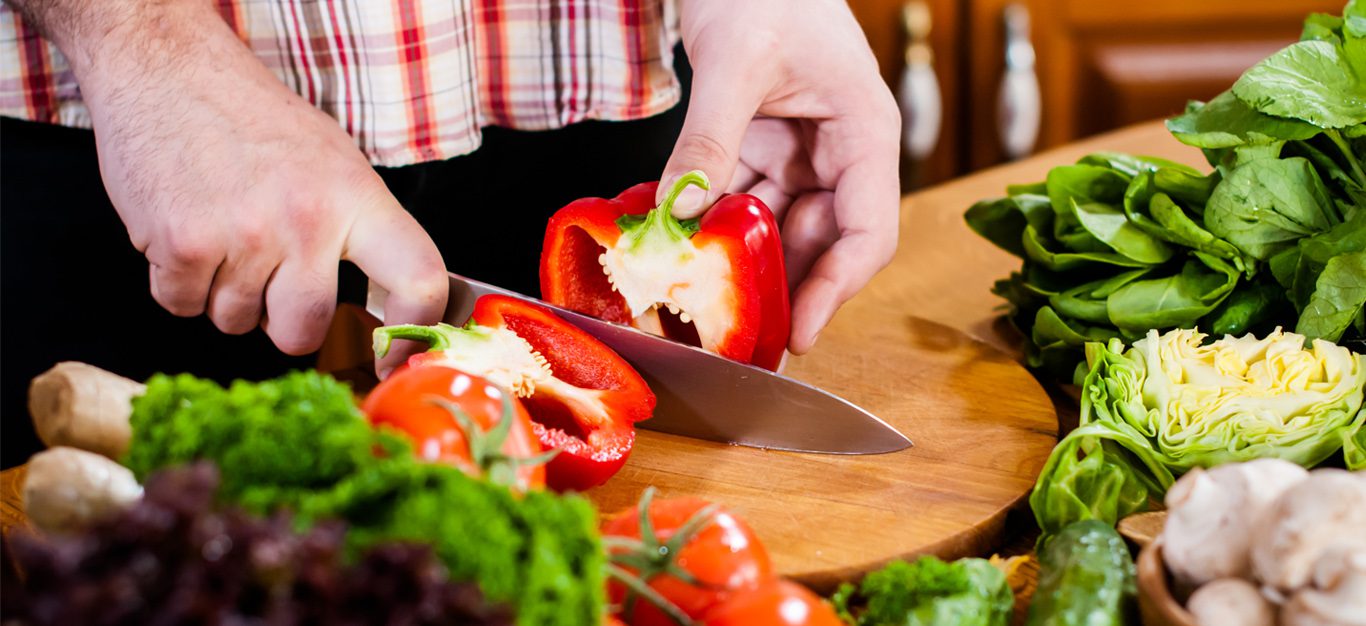 Man's hands holding a knife and chopping a bell pepper on a cutting board, with other vegetables - Plant-based diets have been shown to reduce risks of prostate cancer